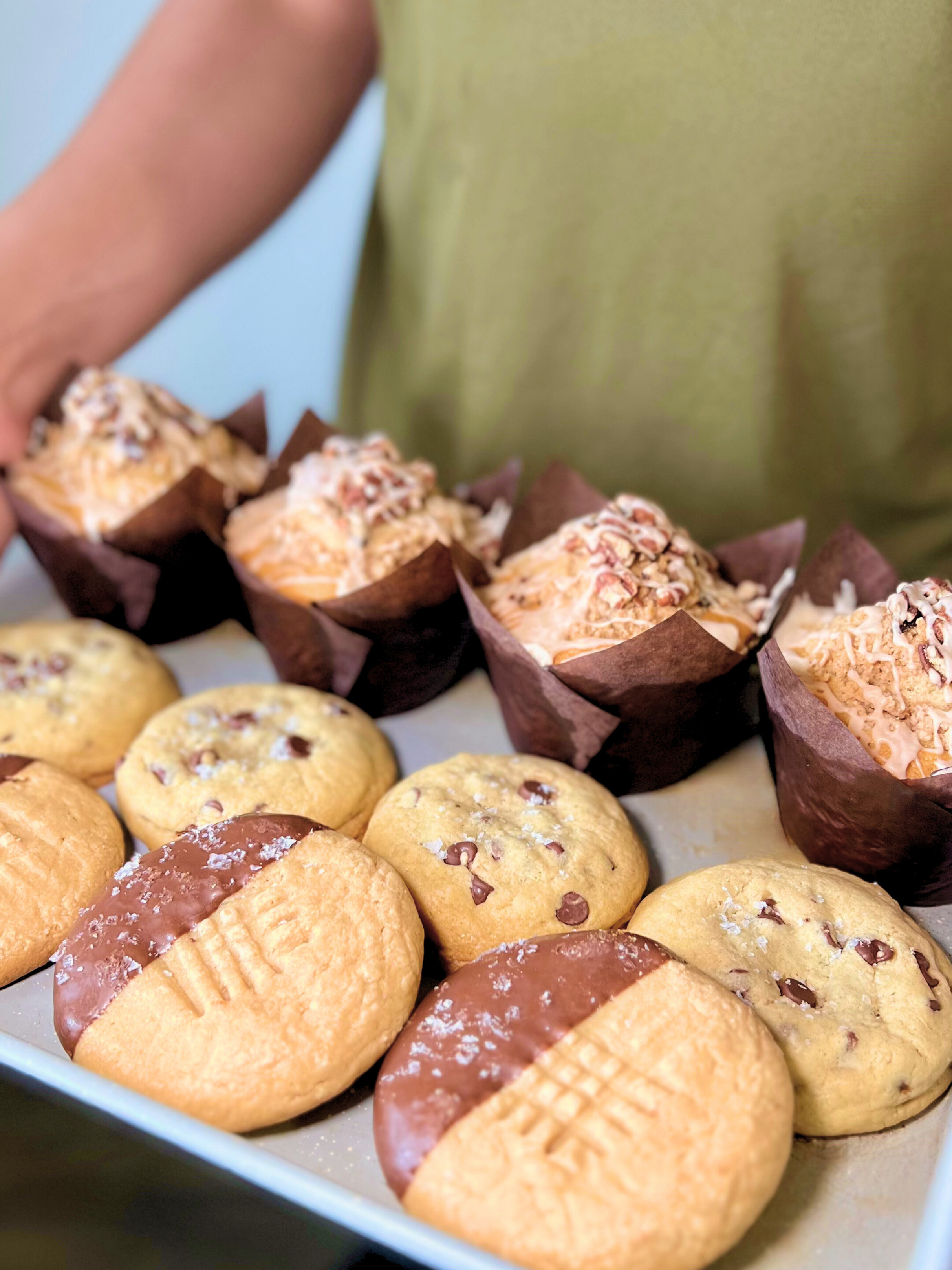 Assorted cookies and muffins on a tray with a person partially visible.