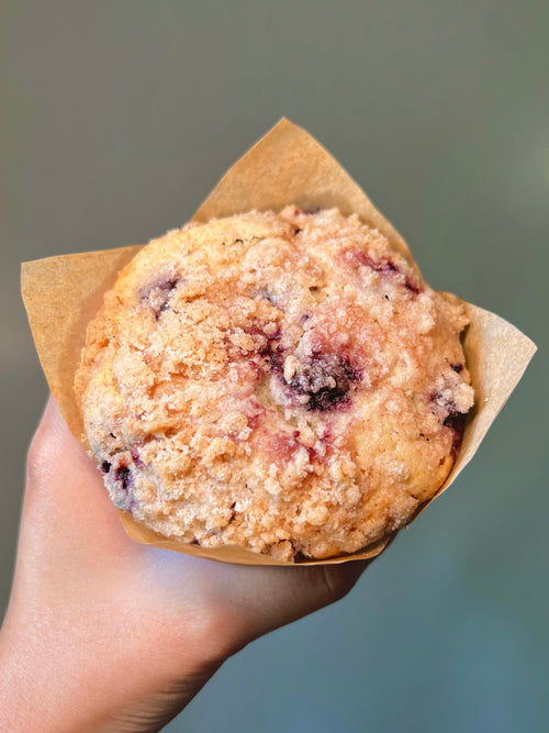Hand holding a Blueberry Crumble  muffin in a brown paper wrapper against a blurred background