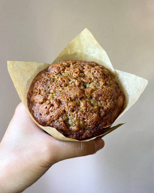 Hand holding a muffin wrapped in brown paper against a neutral background