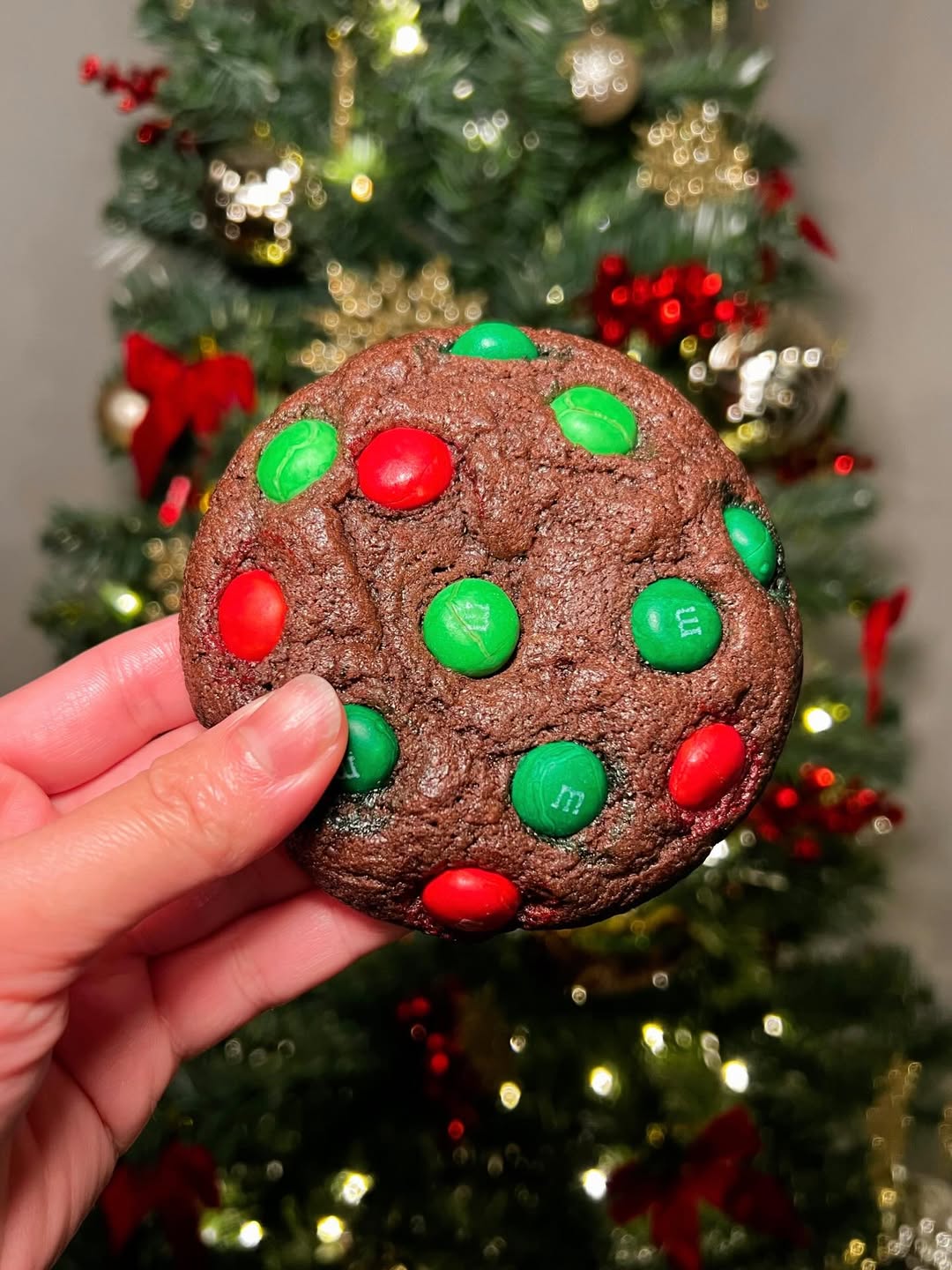 Chocolate cookie with red and green M&M's held in front of a decorated Christmas tree.