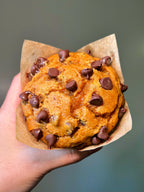 Hand holding a Pumpkin Chocolate Chip Muffin wrapped in brown paper against a blurred background