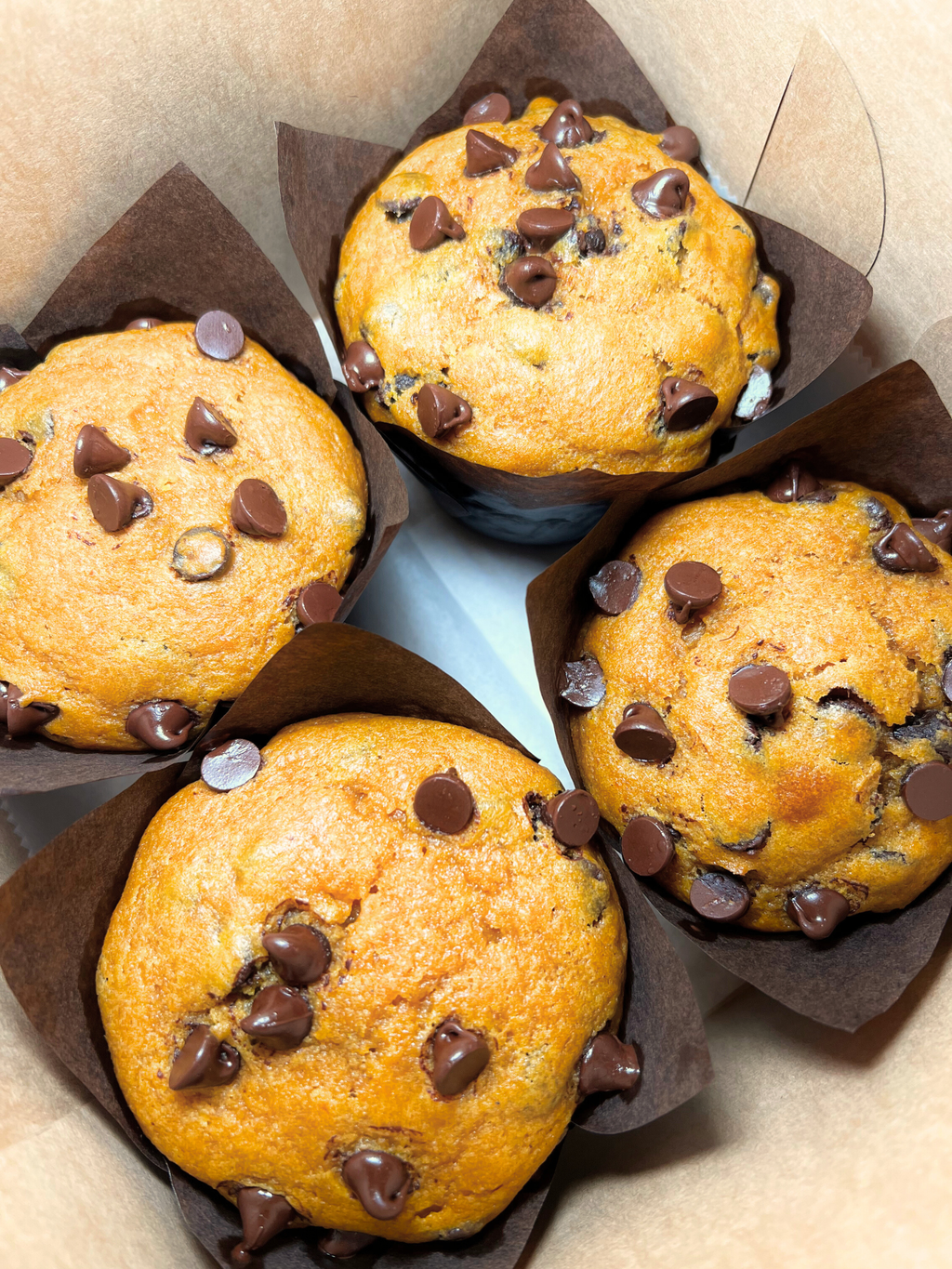 Four Pumpkin Chocolate Chip Muffins in brown paper cups on a light brown background