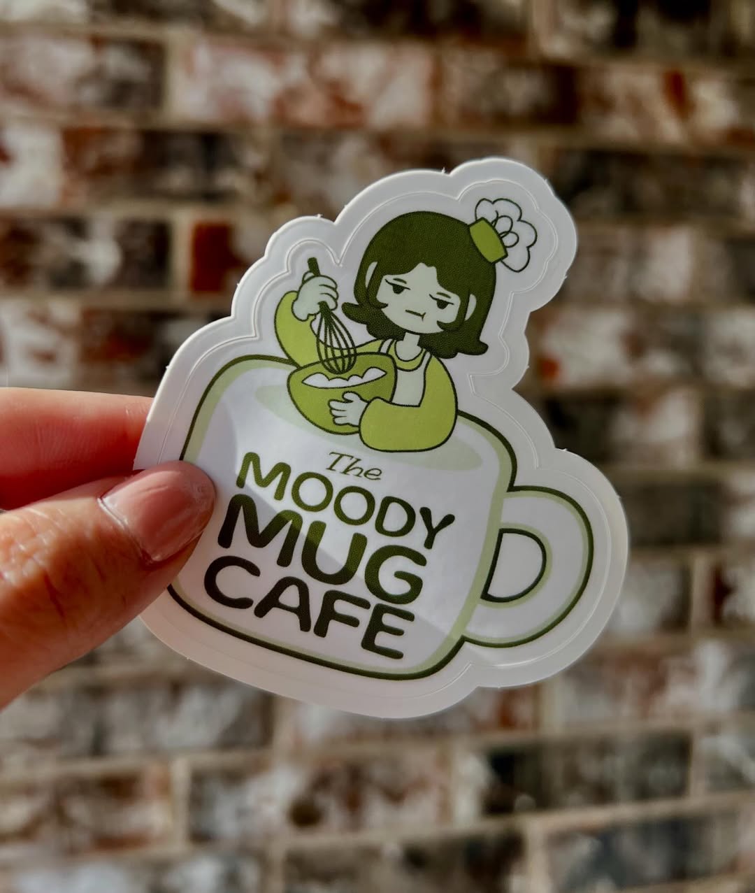 Sticker of The Moody Mug Cafe with a person holding a mug against a brick wall background